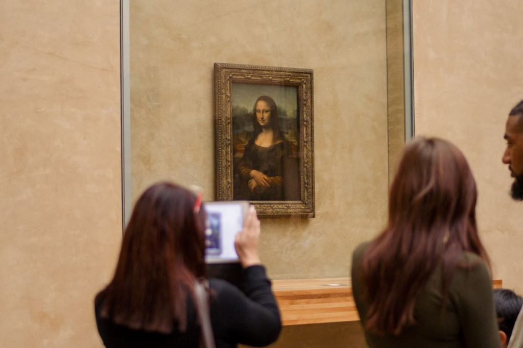 A cfew people stand in the Louvre Museum, Paris, photographing Leonardo da Vinci’s Mona Lisa. Visit as an early bird, or around closing time for the best time to visit the Louvre and see her in person.