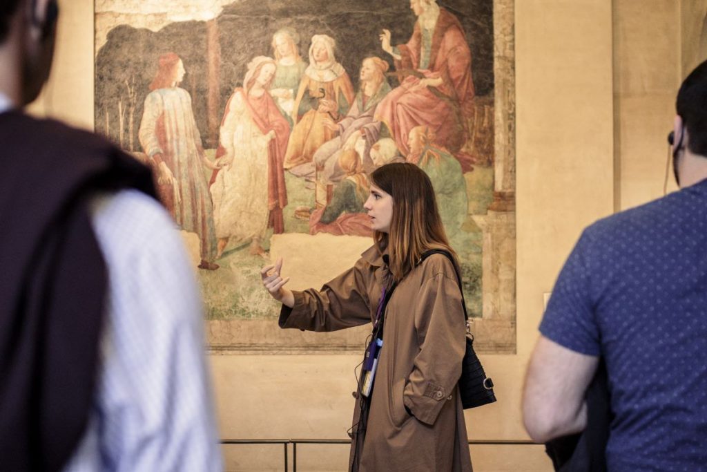 A woman in a brown coat leads a tour at the Louvre, gesturing while explaining a Renaissance mural as two people listen.