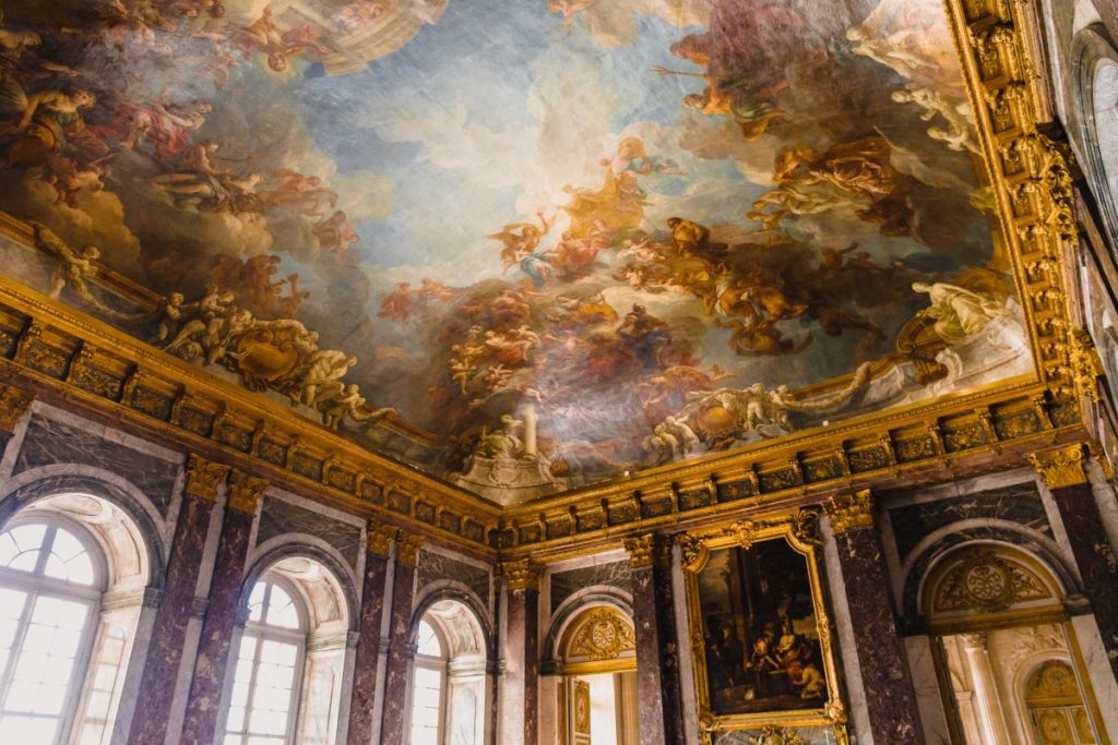 A luxurious hall at the Palace of Versailles features marble walls, gold trim, and a Baroque ceiling painted by Charles Le Brun.