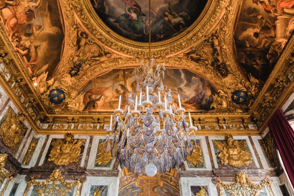 A sparkling chandelier hangs from a gold-accented ceiling adorned with detailed moldings and classical painted murals