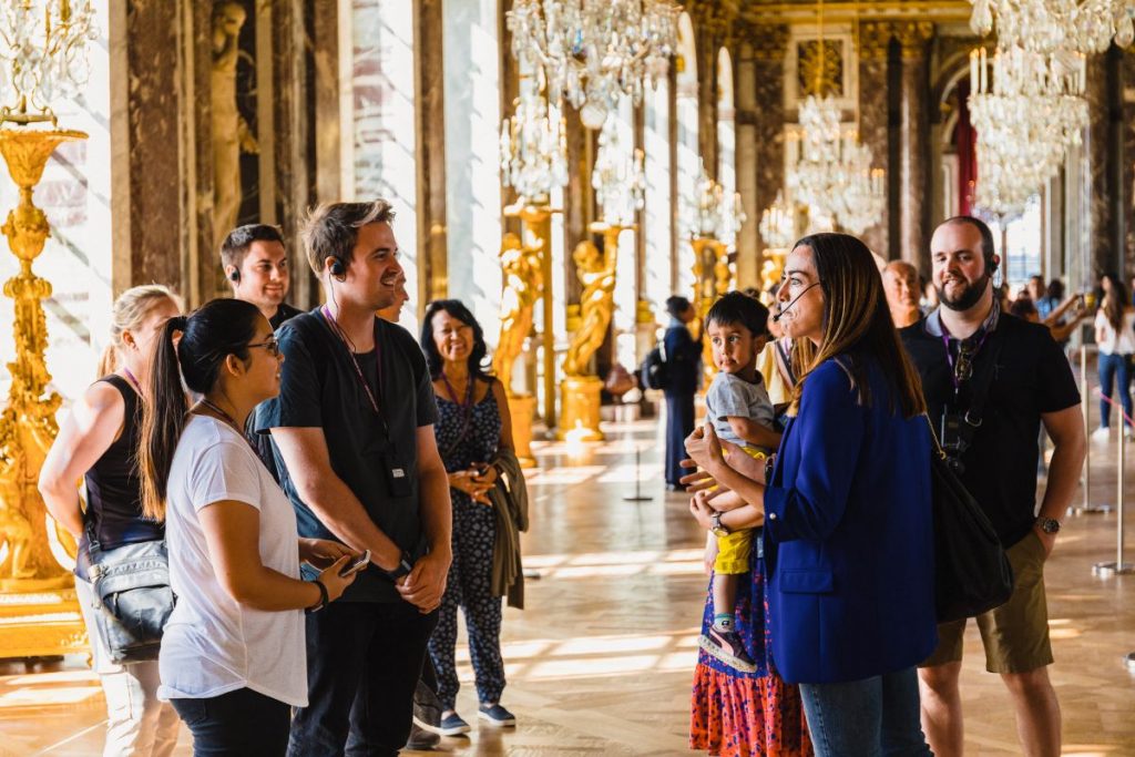 A tour guide explains the Hall of Mirrors at Versailles, surrounded by chandeliers, gilded statues, and tall arched windows.