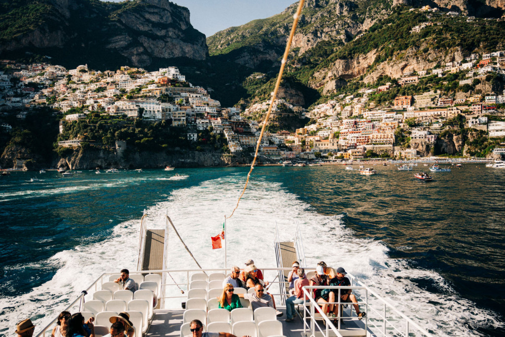 People relax on a boat cruising away from Amalfi Coast, colorful houses and hills behind, water trailing as mountains rise ahead.