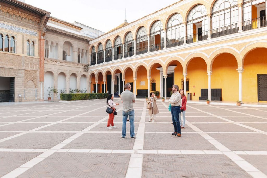 Several people converse in a sunny courtyard with yellow walls, archways, and arched windows at the Alcazar of Seville.
