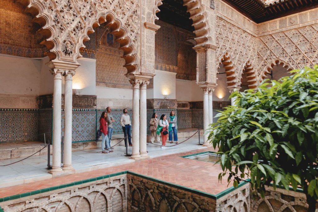 Several people stand beside ornate arches, patterned tiles, and columns in Sevilles Alcazar courtyard - which is why sevilla is worth visiting.
