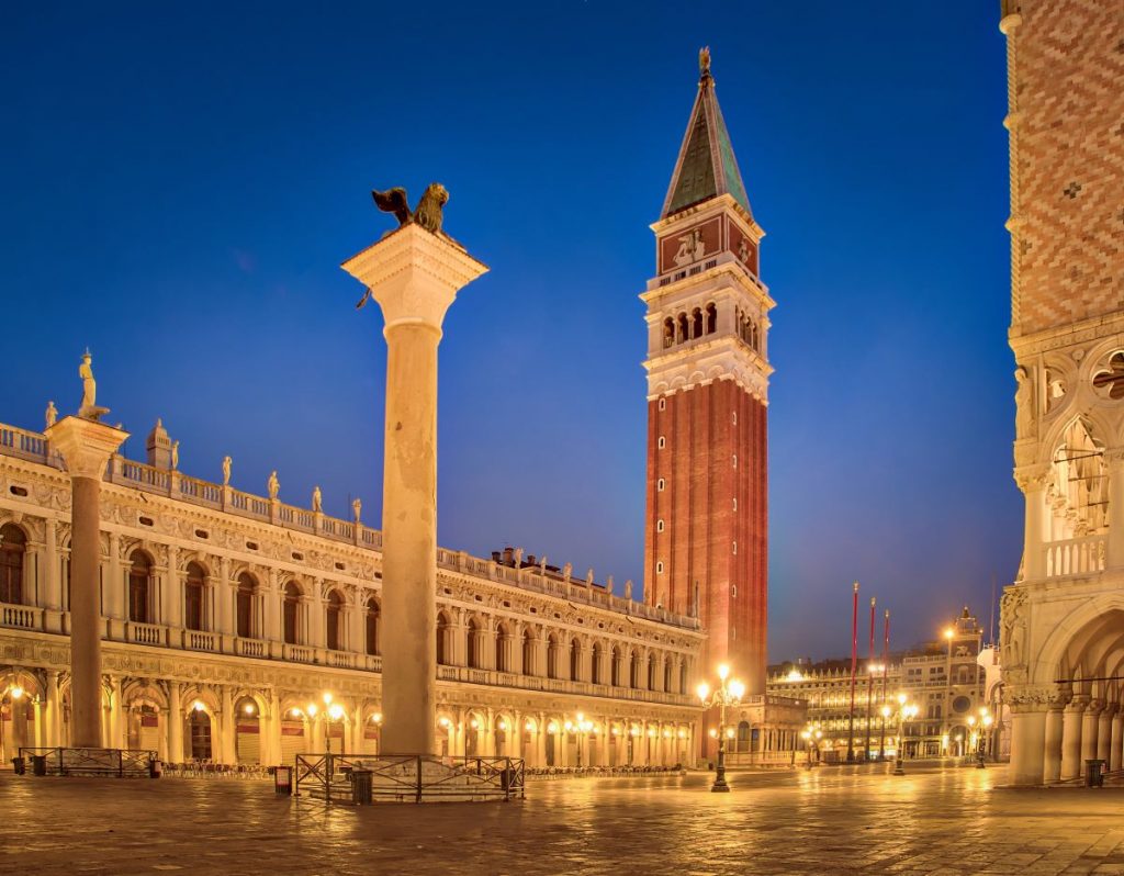 St. Mark’s Square in Venice at night, with the Campanile, winged lion column, and glowing historic buildings under a deep blue sky.