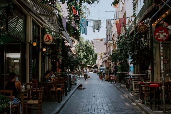 People dine at outdoor cafes along a narrow cobblestone street, with shops, overhead laundry, and lush trees creating a lively scene.