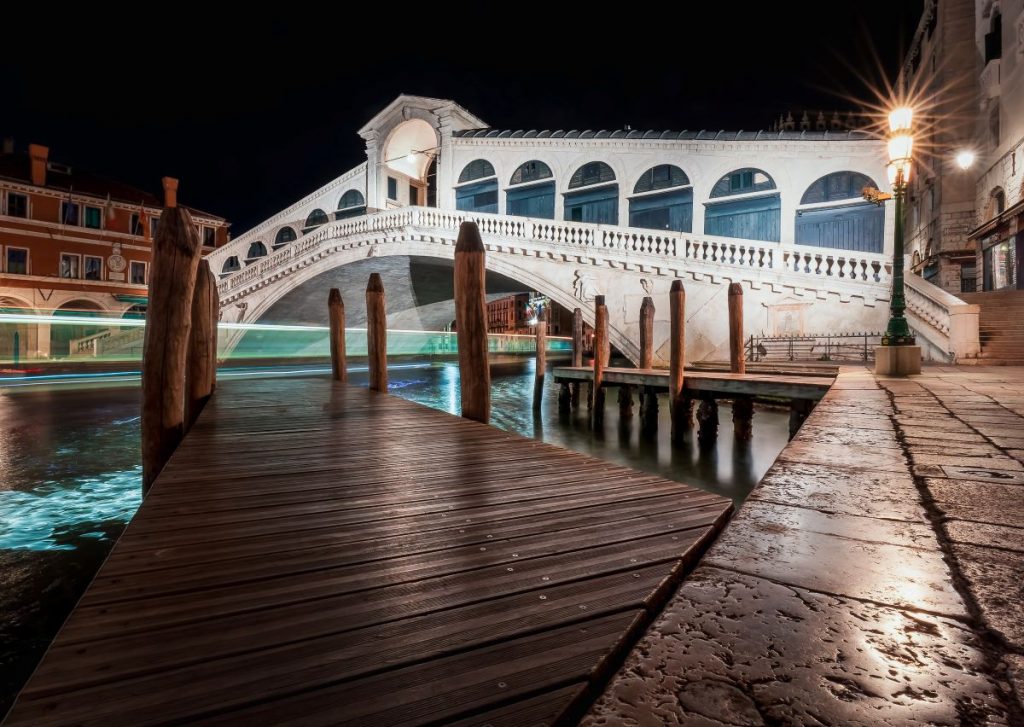 The illuminated Rialto Bridge spans the Grand Canal in Venice at night, with a wooden dock and shimmering boat lights below.
