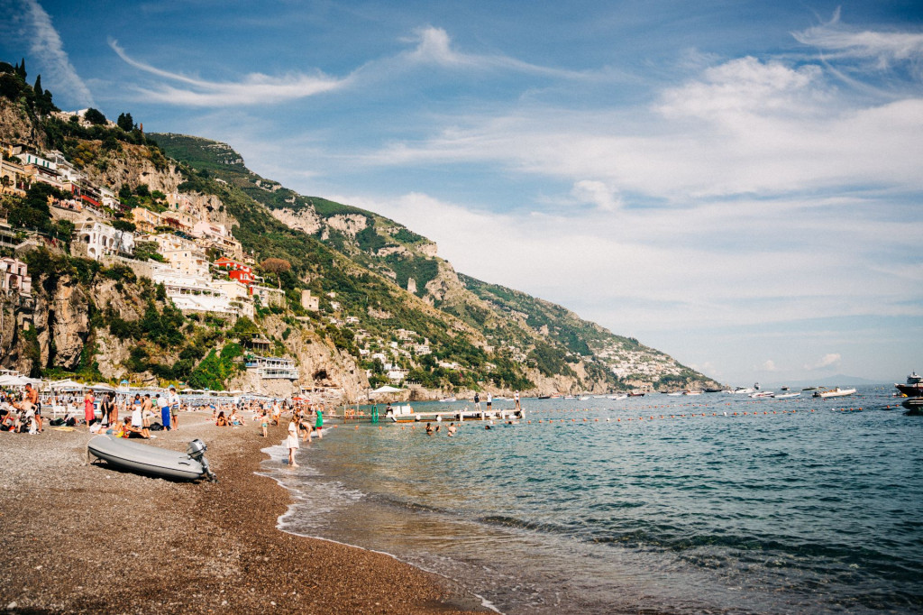 People swim and sunbathe on a lively pebble beach near colorful Amalfi Coast buildings, boats floating beneath green cliffs and blue sky.