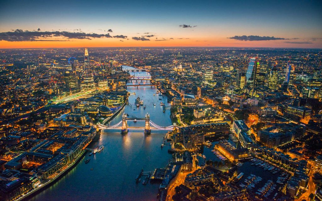 London at dusk, with Tower Bridge lit up and city lights glowing along the Thames as the sky fades from orange to blue.