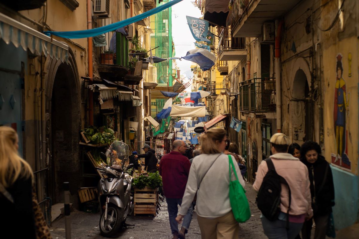 Crowds walk past market stalls and hanging laundry on Via San Gregorio Armeno in Naples, surrounded by colorful old buildings.