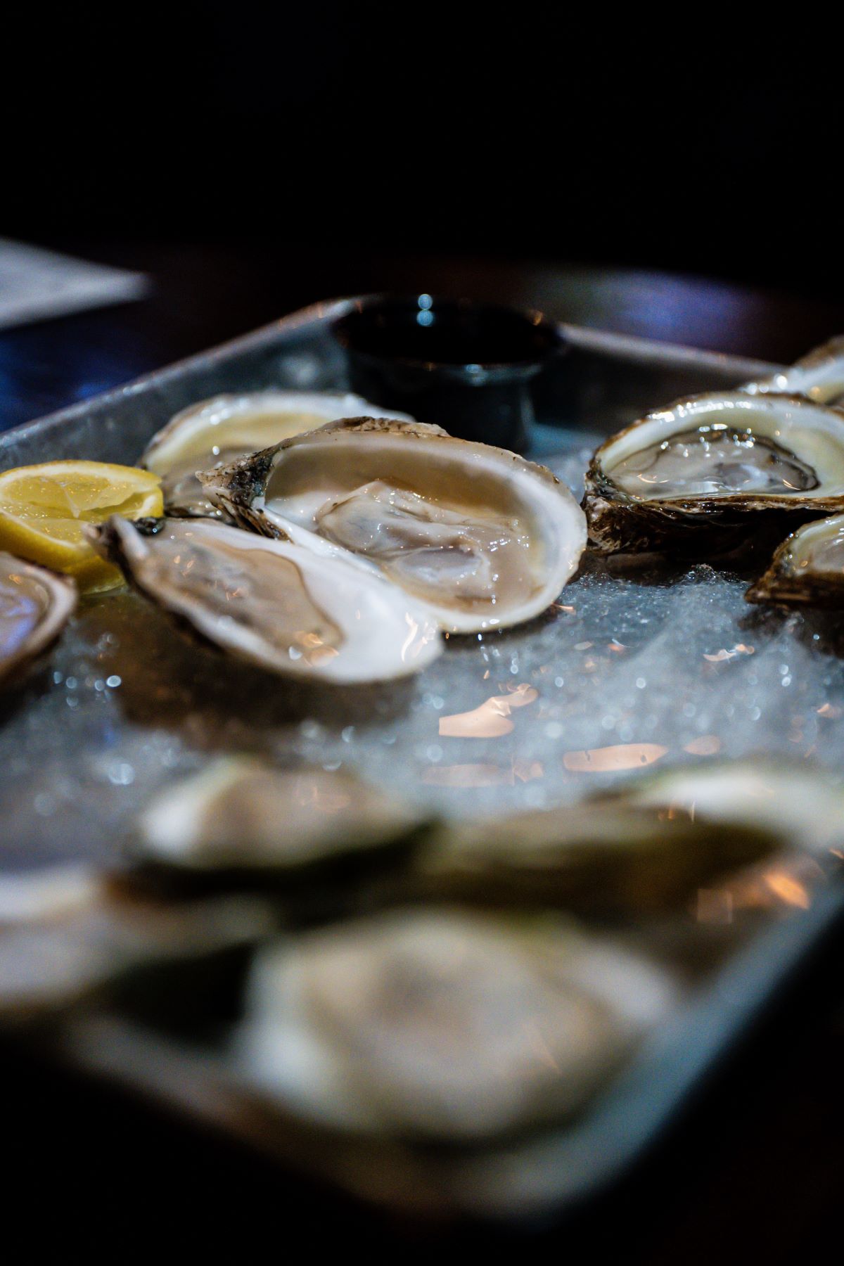 Freshly shucked oysters rest on ice with lemon wedges and a cup of dipping sauce on a metal tray, ready to eat.