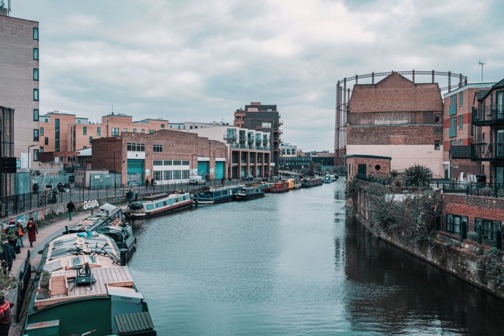 A narrow canal in London features houseboats, brick buildings, lively oyster bars, pedestrians, and a large cylindrical gas holder structure.