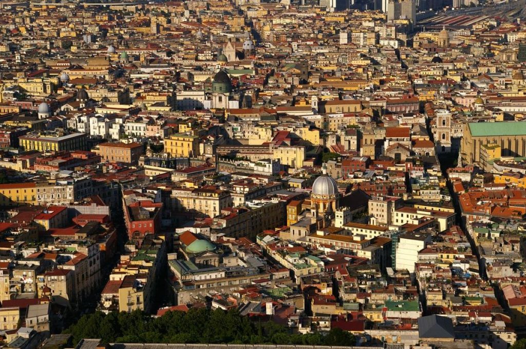 Aerial view of Naples, Italy with vibrant buildings, historic domed churches, and winding streets illuminated by bright sunlight.