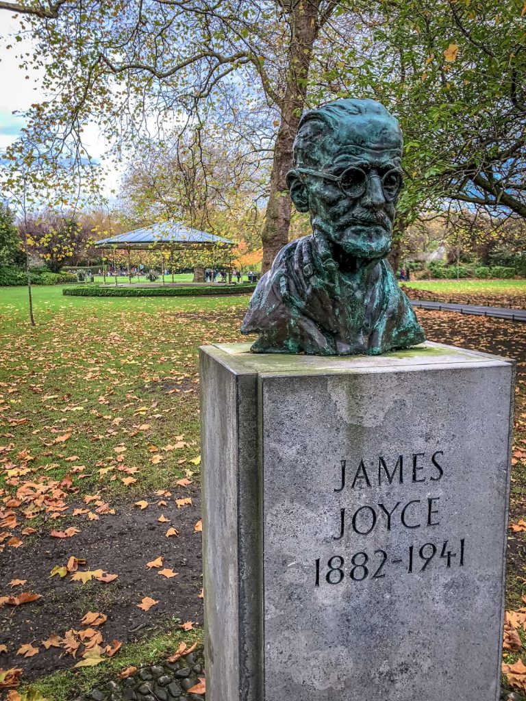 A bronze bust of James Joyce on a stone pedestal stands in a leafy Dublin park, honoring the famed Irish writer.