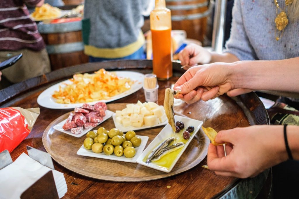 A person in Spain lifts a pickled fish with a fork, surrounded by plates of olives, cheese, salami, and chips.