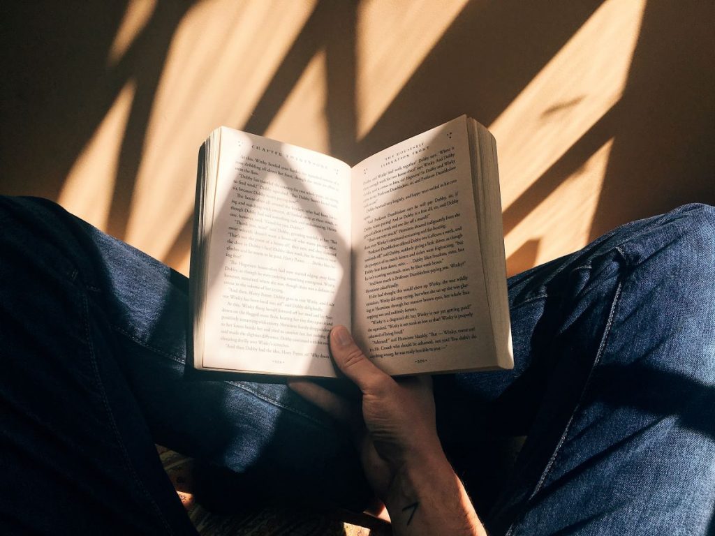 A person wearing blue jeans sits cross-legged holding an open book in their lap, sunlight casting warm diagonal shadows.