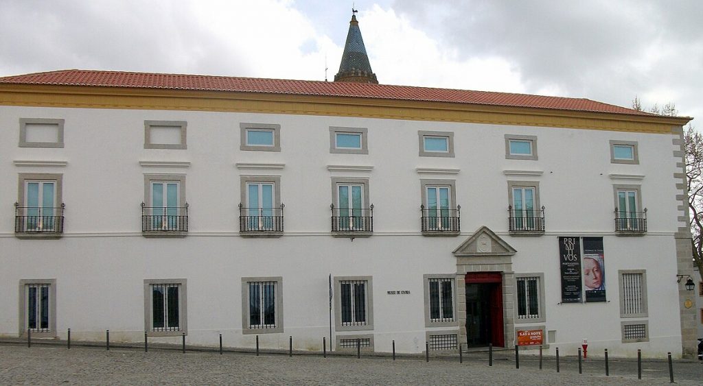 A white, two-story building with a red-tiled roof stands on a cobblestone street, the Museu de Evora is one of the things to do in Évora, Portugal; church tower visible.