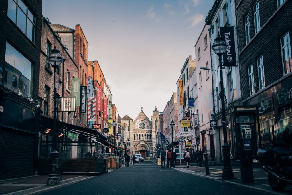 Pedestrians walk past colorful shops and a famous Dublin bookstore, with a large historic stone church at the street’s end.