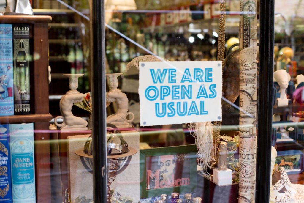 A bookstore in Dublin displays a blue We are open as usual sign, with neatly arranged books, decor, and figurines visible inside.