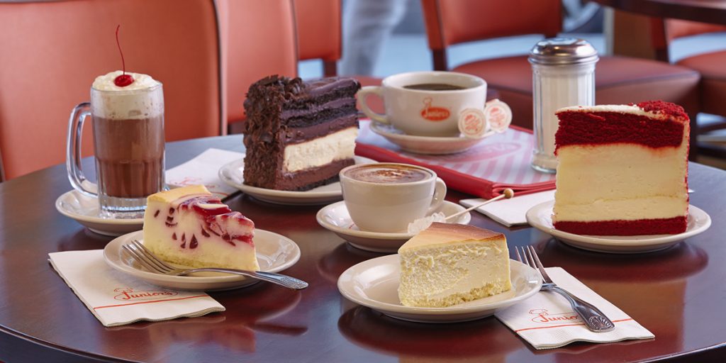 Slices of cheesecake, red velvet cake, and chocolate cake sit on a round table with coffee drinks in a cozy café recommended on the Brooklyn Food tour 