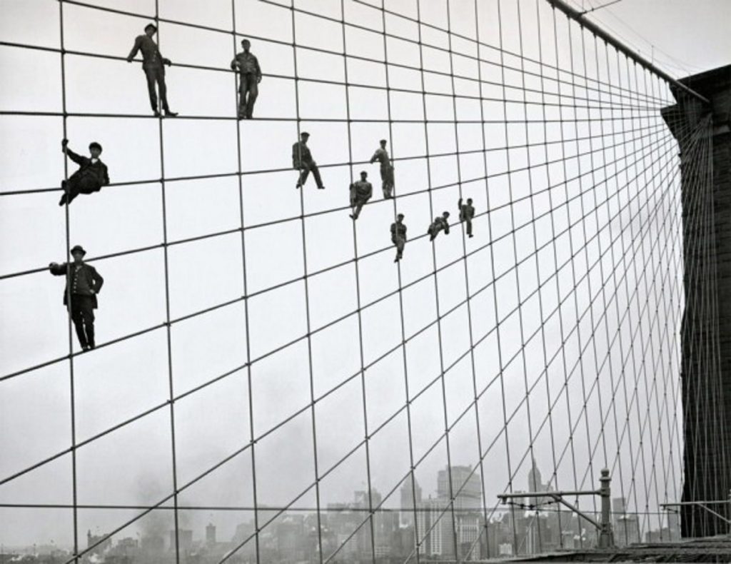 Men climb and balance on the suspension cables of Brooklyn Bridge high above New York City, photographed in dramatic black and white.