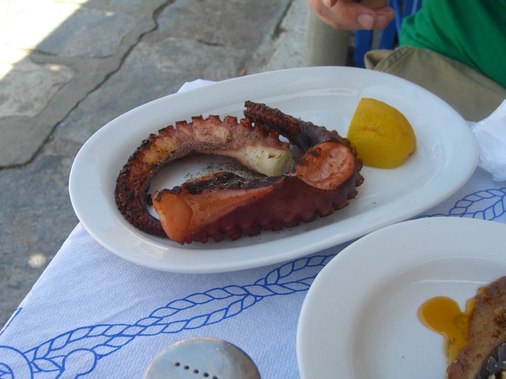 A cooked octopus tentacle rests on a white plate with a lemon wedge, atop a blue-patterned cloth in Plaka, Athens.