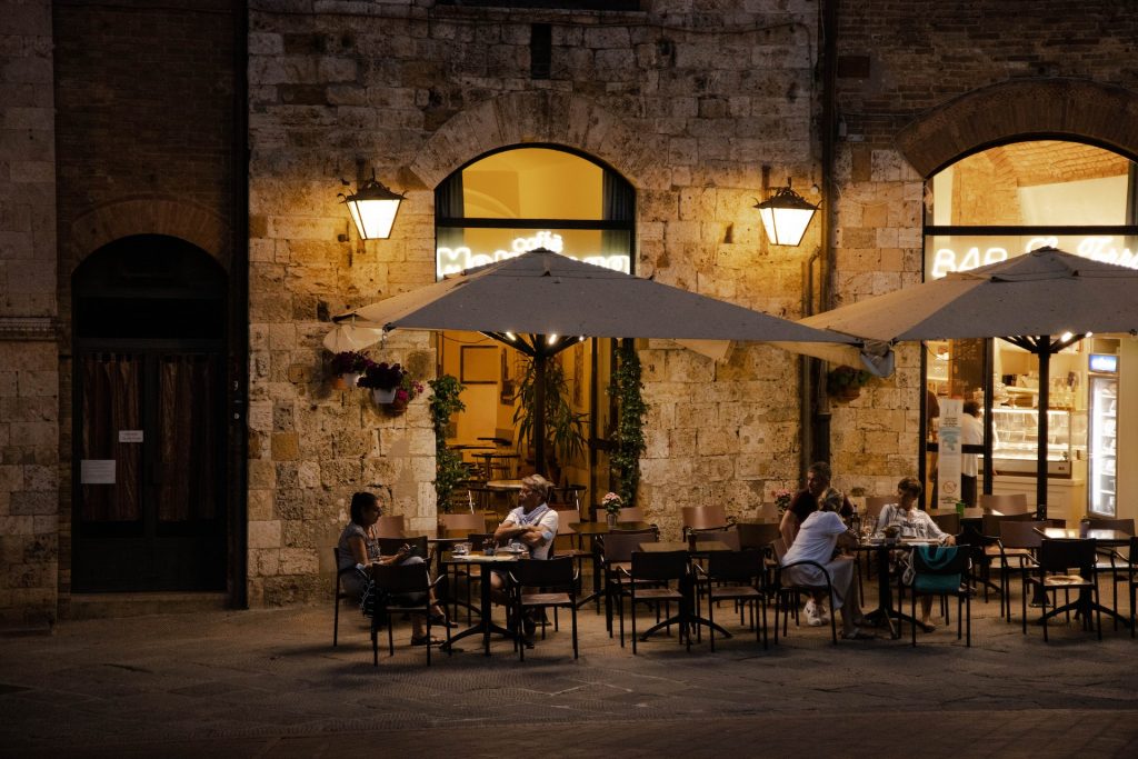 People enjoy drinks at a Siena street café at dusk, sitting under umbrellas beside warmly lit stone buildings on cobblestones.