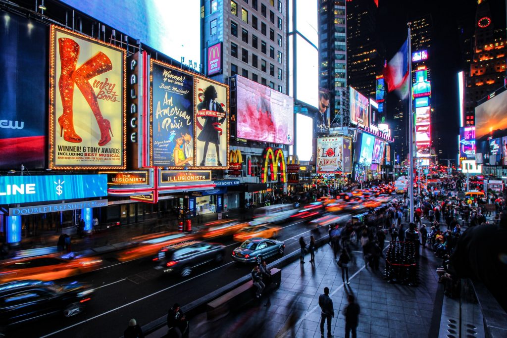 Times Square in New York City glows at night with flashing billboards, bustling crowds, and streaks of headlights from passing cars.