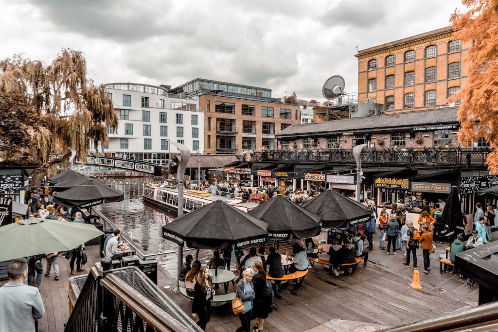 A lot of people eating outside on a boardwalk with tables and umbrellas, at the open air Camden Market during a rainy day