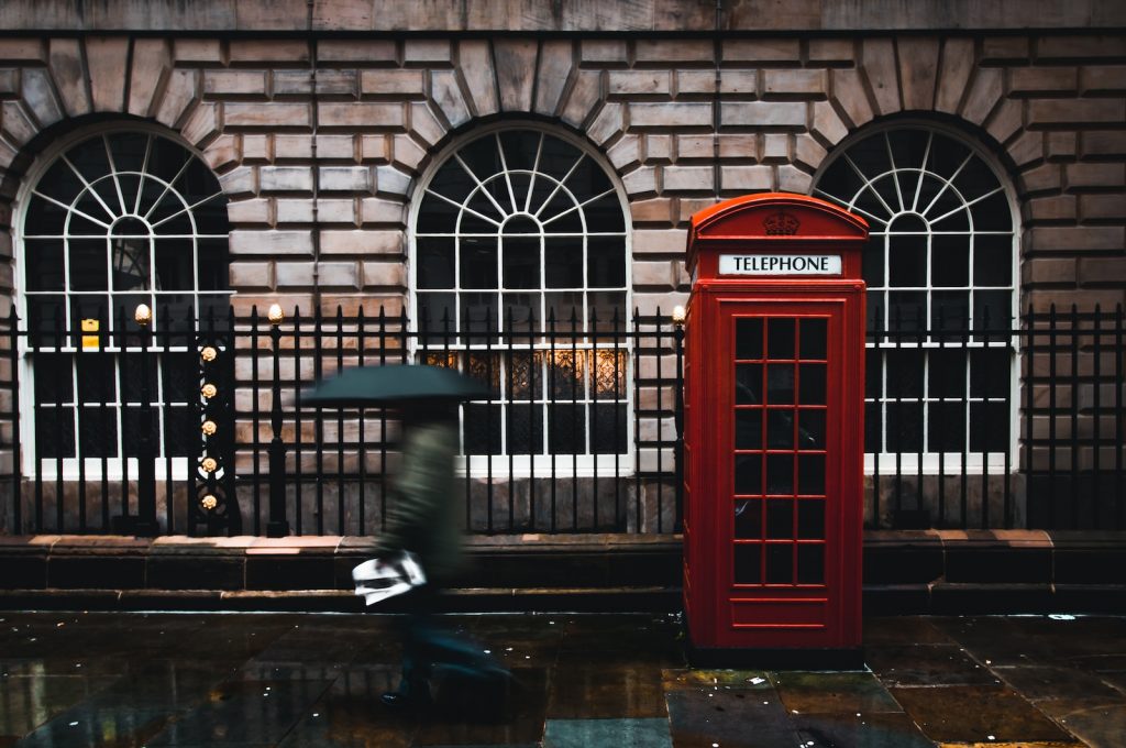 A person carrying an umbrella walks past a red British telephone booth on a rainy London street near arched stone buildings.