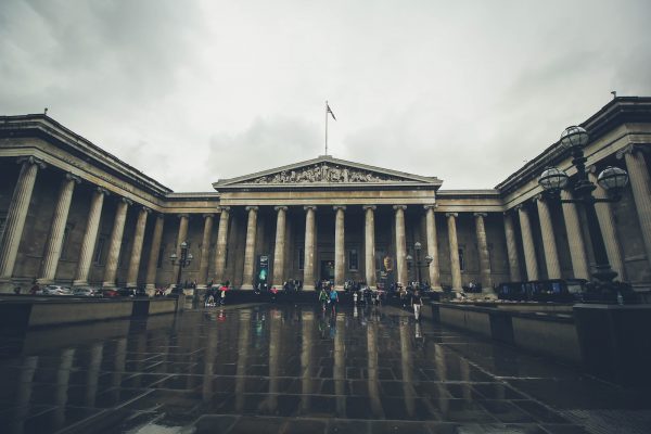 Several people walk across a wet, reflective plaza toward the British Museum’s grand neoclassical entrance on a cloudy day.