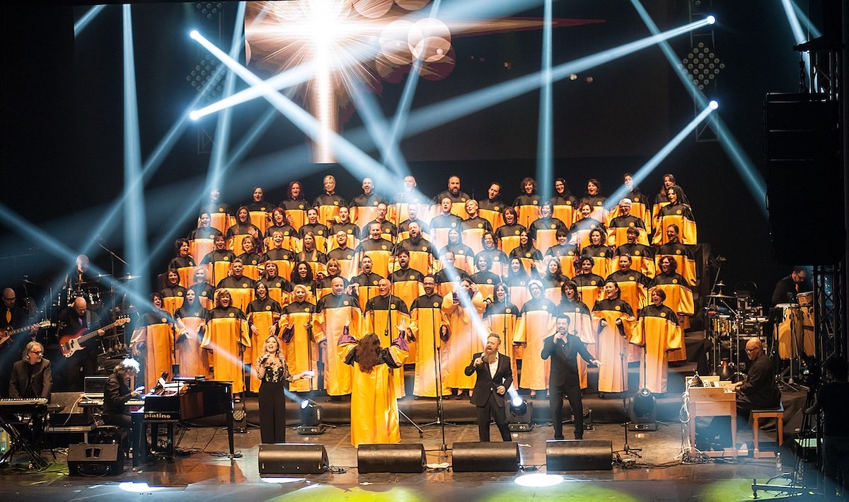 A large Choir wearing yellow and black robe, performing on a stage with bright spotlights and live musicians