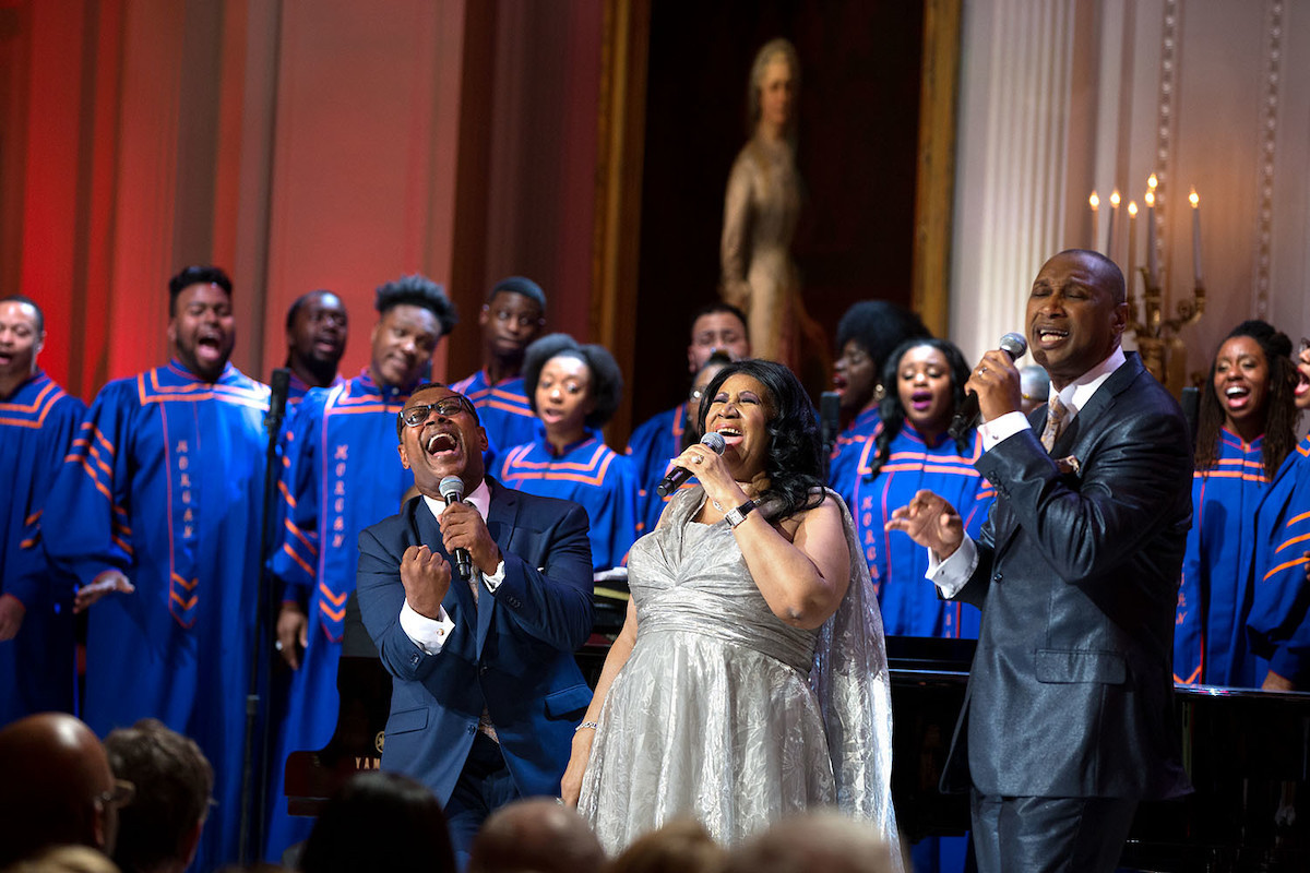 Aretha Franklin performing with 2 male singers, and backing singers in blue and orange robes behind them.
