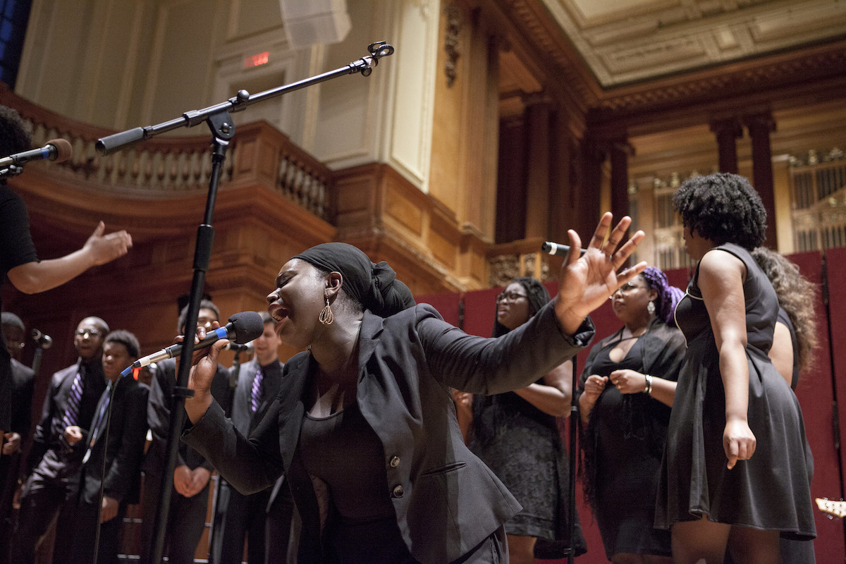 A women sings passionately into a microphone with other choir members all dress in black behind her, in a church setting.