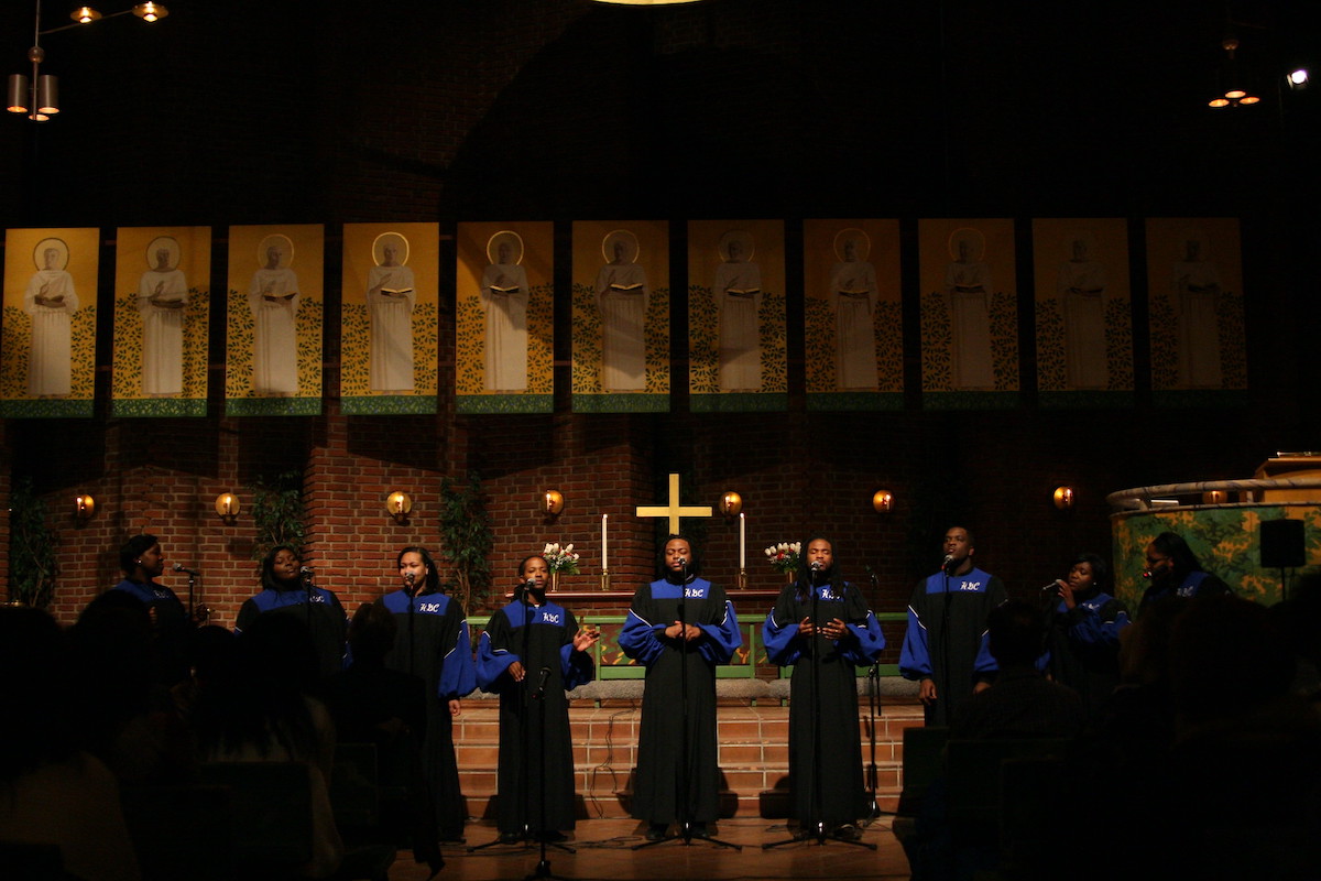 The Howard Gospel Choir in black and blue robes, performing inside a church, facing the congregation, with the cross and religious paintings behind them
