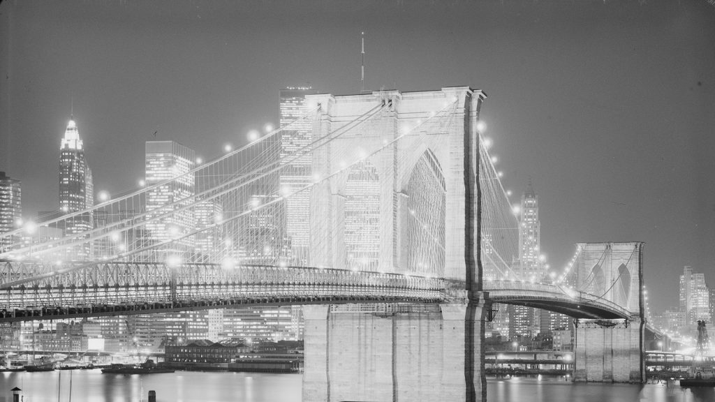 People walk across the illuminated Brooklyn Bridge at night, with New York City's glowing skyline and skyscrapers reflected in the river.