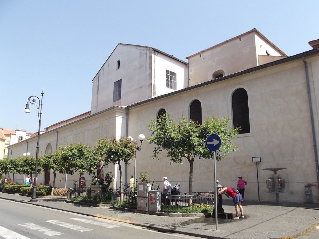 a street scene in sorrento Italy. with a white building with arched windows, and a sidewalk with olive trees and a person drinking from water fountain