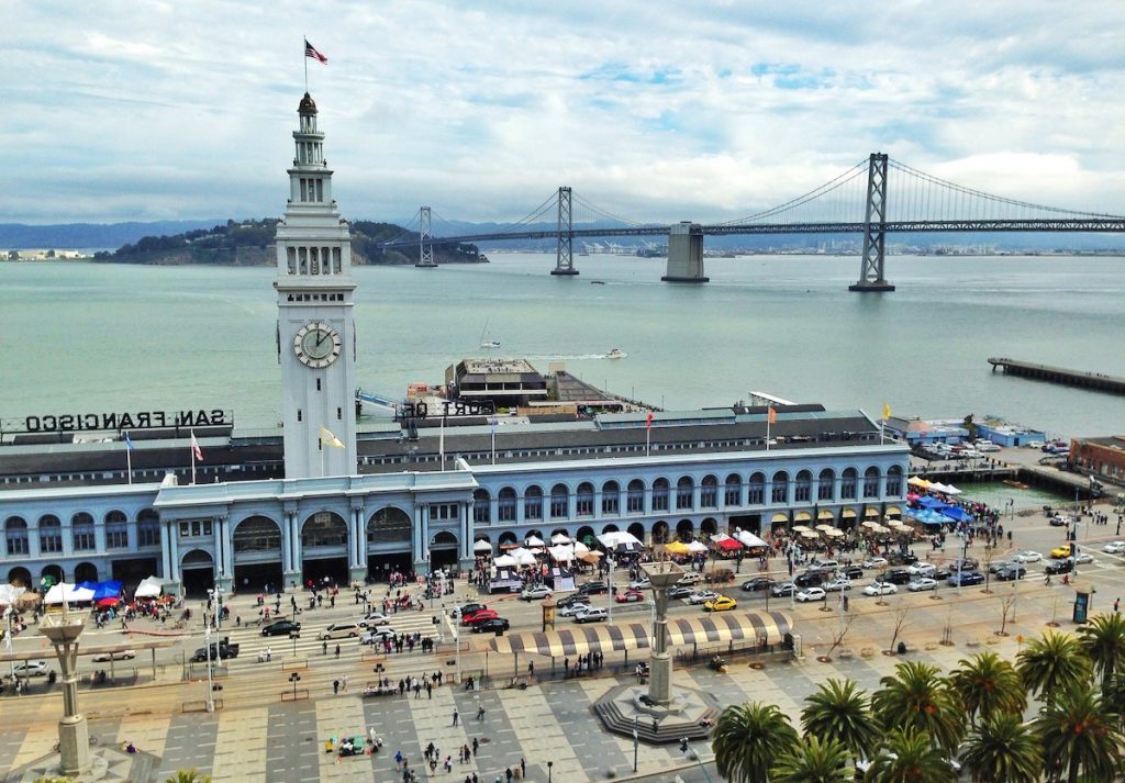 The Ferry Building’s clock tower rises above bustling outdoor market crowds by the waterfront, with the Bay Bridge in the background.