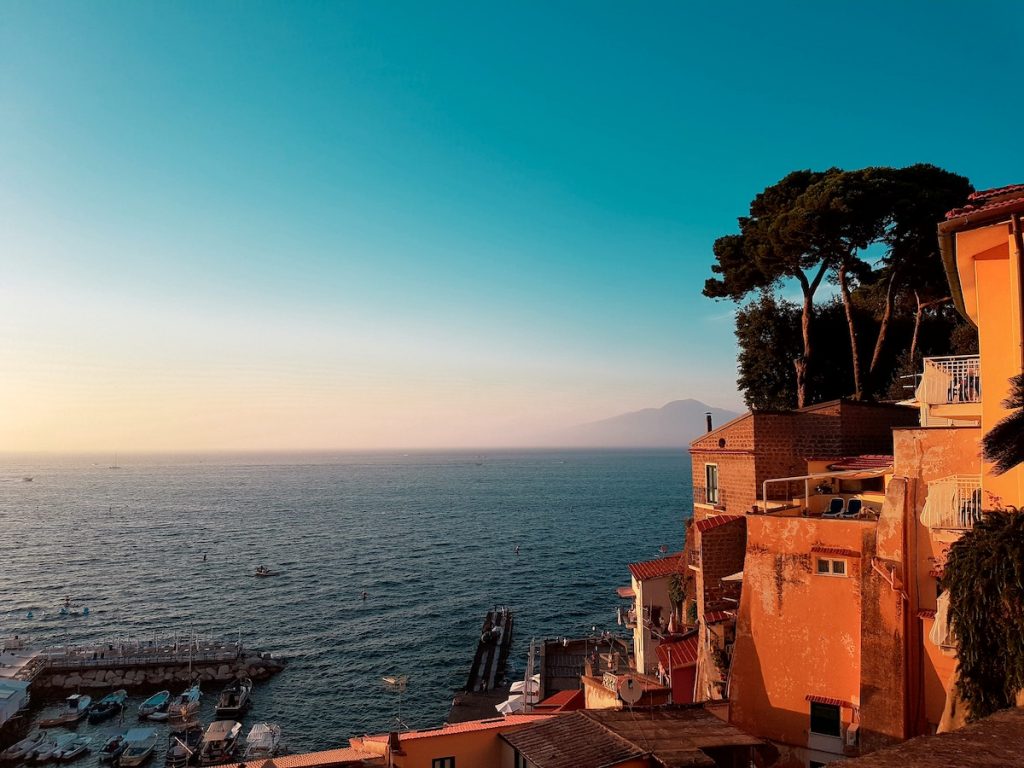 Colorful hillside buildings in Sorrento overlook a marina and calm blue sea, with distant hazy mountains beneath a clear sky.