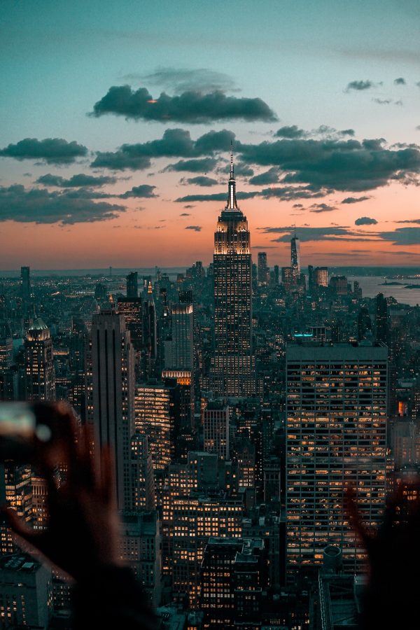 At sunset in New York City, silhouetted hands hold phones to photograph the illuminated Empire State Building among surrounding skyscrapers.
