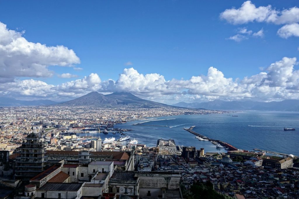 Naples, Italy stretches across the foreground with colorful buildings, a busy harbor, and Mount Vesuvius towering under a clear sky.