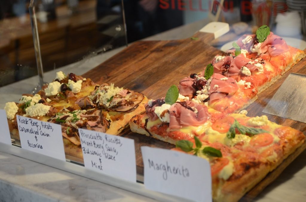 Three types of rectangular traditional Italian pizza with various toppings sit on a wooden board behind glass at a pizzeria counter.