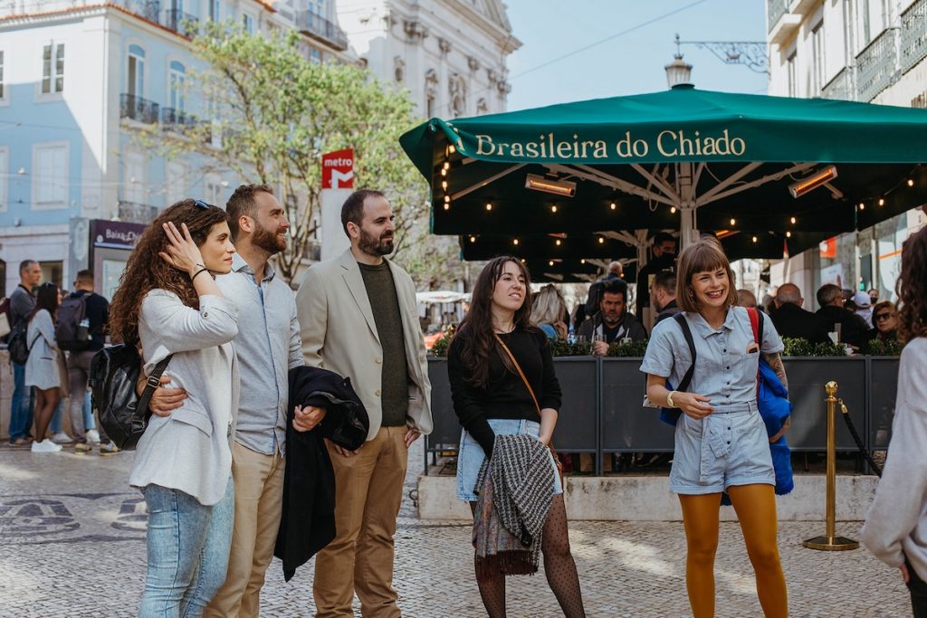 Five people on a tour smile together in Lisbon’s bustling Chiado plaza near Café A Brasileira, with classic buildings and a metro sign behind them. Lisbon is one of the best places for a solo trip to Portugal.