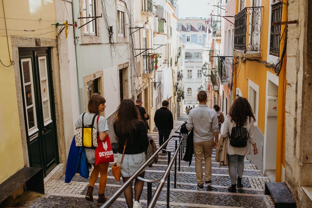 Several people stroll along a narrow cobblestone street with colorful buildings and balconies, approaching a sunlit plaza during a food and fado in Lisbon Tour 