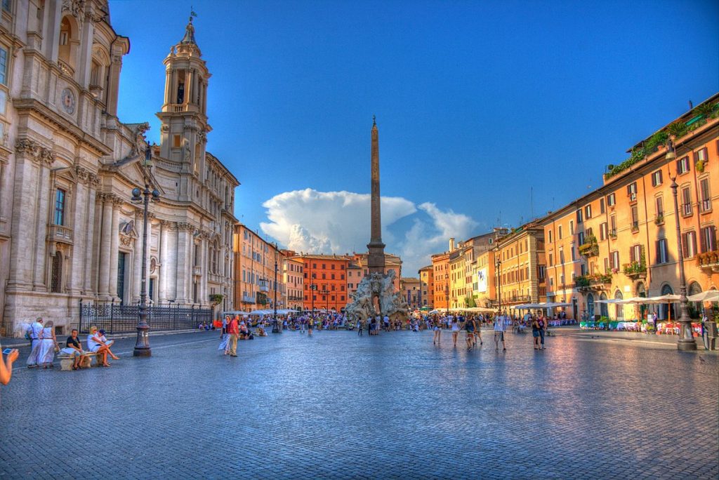 Crowds walk and relax in Rome’s Piazza Navona, surrounded by historic buildings, SantAgnese in Agone church, and the ancient obelisk.