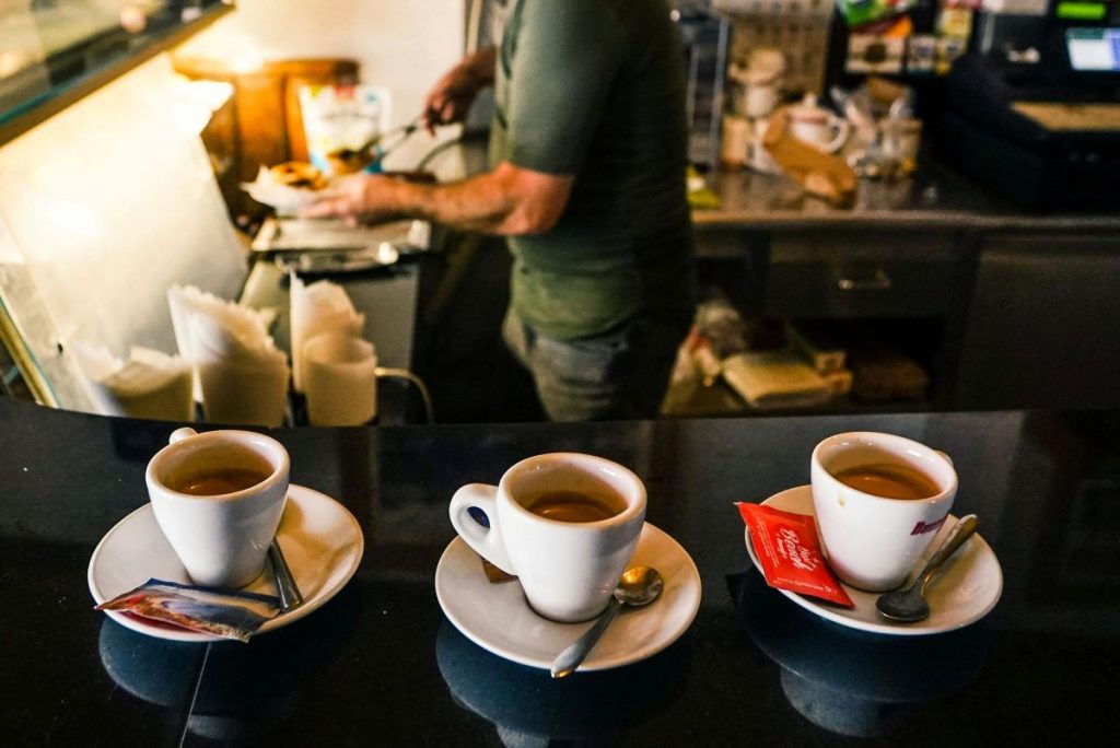 Three cups of espresso on saucers with sugar packets sit on a counter in an Italian café as a barista prepares food.