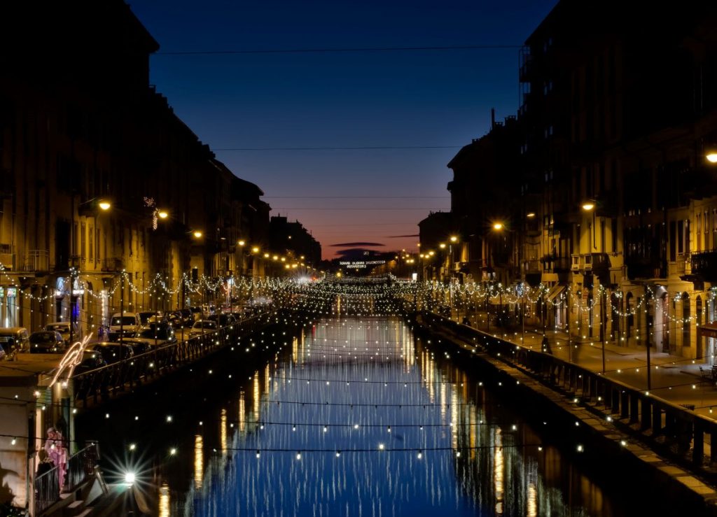 Navigli is worth visiting when in Milan, for a night our on the town. A canal flanked by italian architecture, with string lights over the water, and a view of the sunsetting at dusk in the distance