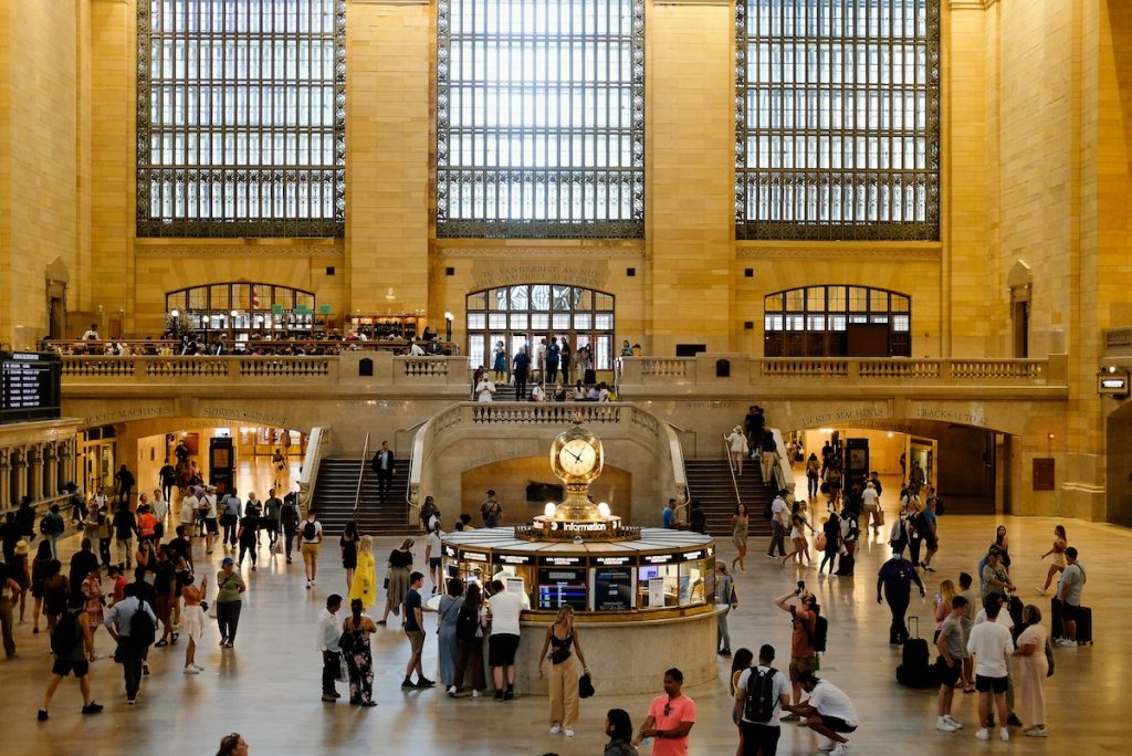 Crowds move past the iconic four-faced clock atop the information booth in Grand Central Terminal, sunlight streaming through arched windows.