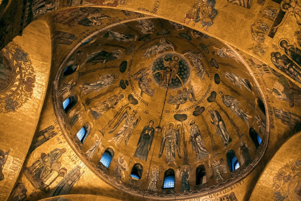 A golden mosaic dome at St. Mark’s Basilica displays saints, biblical scenes, and Latin inscriptions, glowing in natural sunlight.