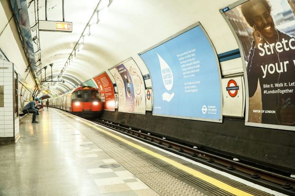 A red London Underground train approaches Oxford Circus station. Commuters wait near bright ads, including one with a smiling person stretching.